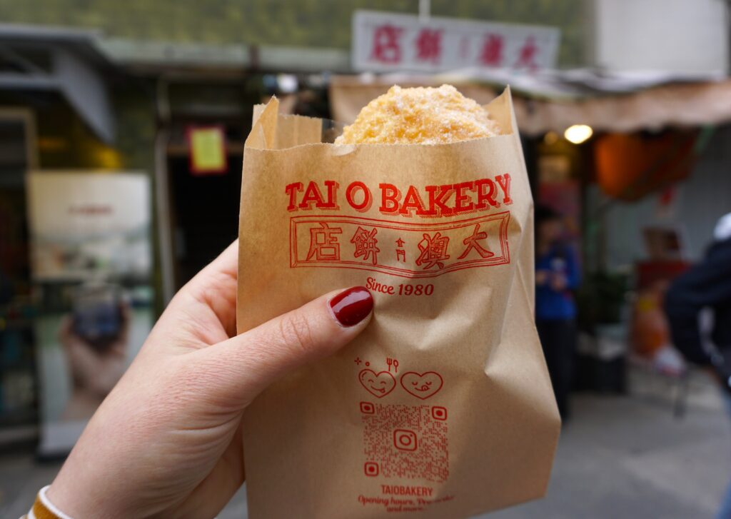 Hand with red nail polish holding a sugar covered Tai O Doughnut in a brown paper bag with red letters that read, "Tai O Bakery sing 1980."