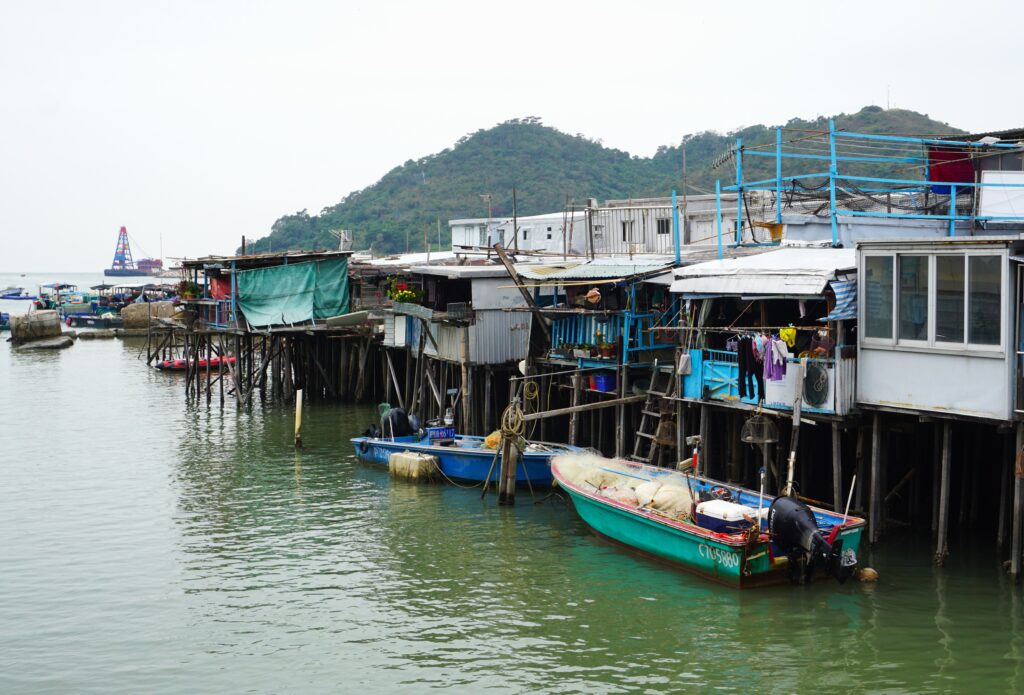 Row of wooden stilt houses and small fishing boats along the waterfront in Tai O, with a hill and other boats visible in the background.