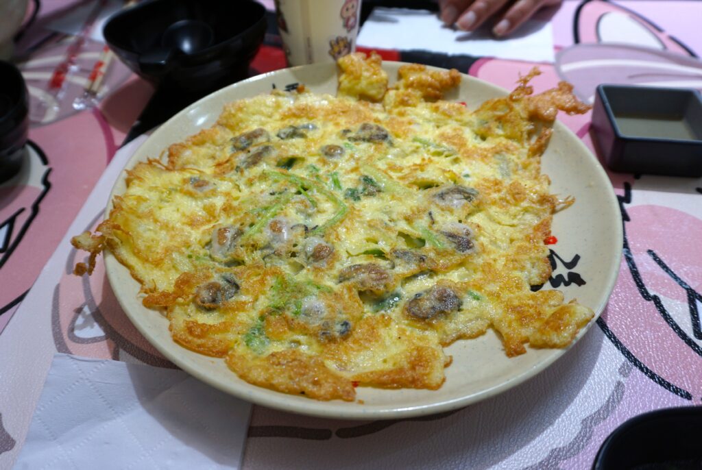 The image shows a savory dish served on a round, beige plate. The dish appears to be a pancake or omelet with a crispy, golden-brown crust on the edges. It contains visible ingredients such as small pieces of seafood and sliced green vegetables scattered throughout. The plate sits on a pink and white tablecloth with illustrations. Surrounding the plate are various dining items, including a small square dipping dish, a pair of chopsticks in a wrapper, and a black bowl. In the background, a hand is visible near a glass containing a beverage.
