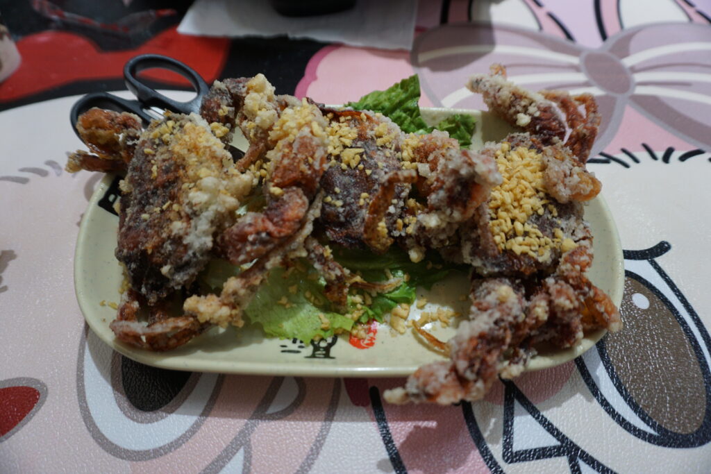 The image displays a dish of fried crabs placed on a rectangular, cream-colored plate. The crabs are coated with a layer of batter and sprinkled with small, chopped nuts. They sit on a bed of green lettuce leaves. On the top left corner of the plate, a black pair of scissors is partially visible. The plate rests on a tabletop featuring a colorful, abstract pink and red pattern, with parts resembling cartoon eyes and hair bows.