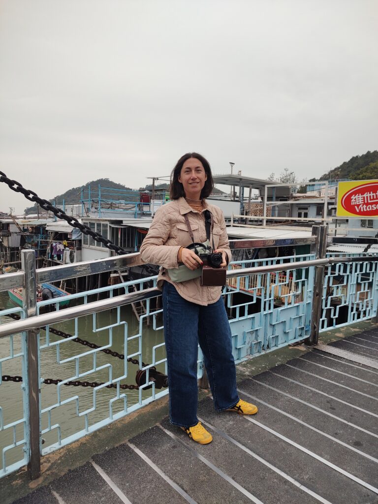 The author, Tamar, stands on a metal bridge in Tai O fishing village, holding a camera, with boats and buildings on water in the background under a cloudy sky.