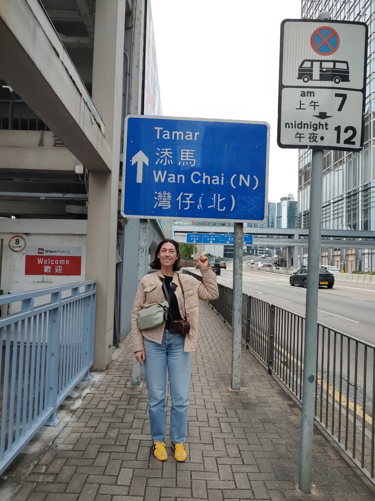The author standing on a sidewalk in Hong Kong pointing up at a blue road sign that reads Tamar and Wan Chai North with Chinese characters and an upward arrow. Urban street scene with tall buildings cars and pedestrian railings in the background highlighting directions in the city.