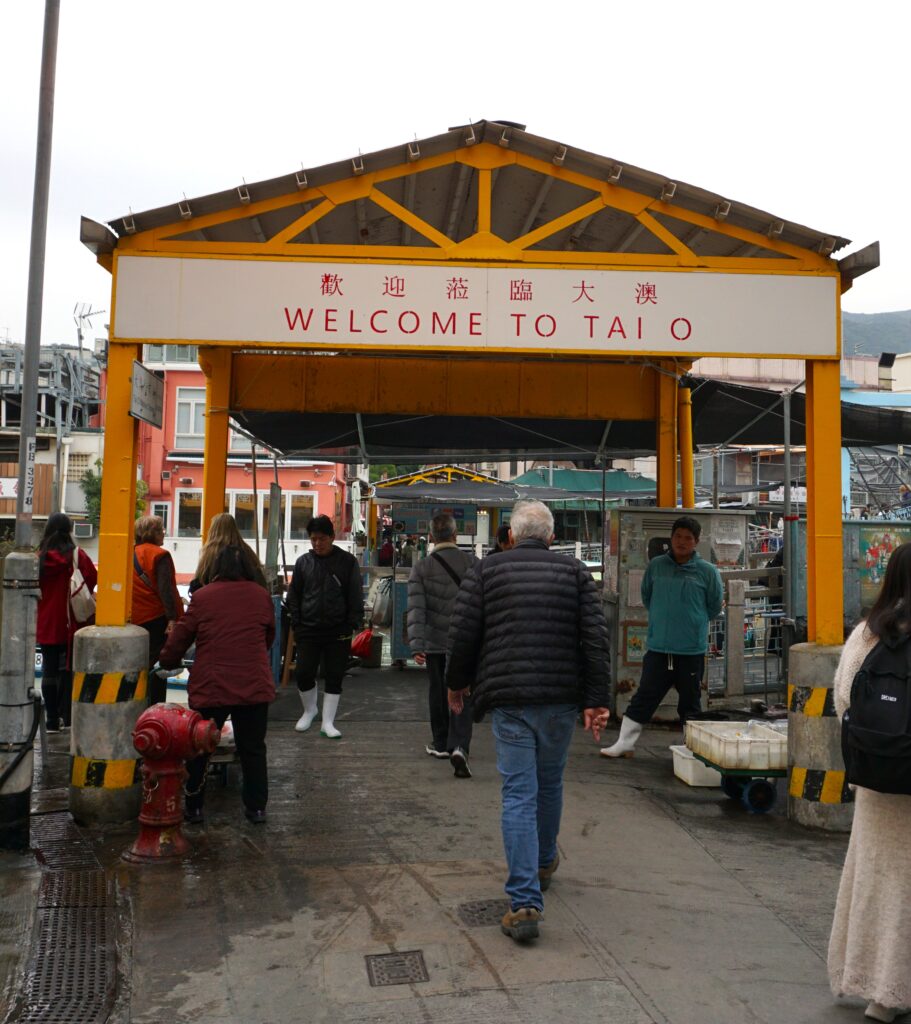 People walk beneath a yellow entrance arch that reads Welcome to Tai O with Chinese characters above it. The covered walkway leads into Tai O fishing village in Hong Kong with market stalls signage and daily activity visible in the background.